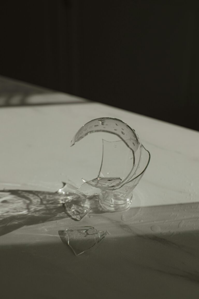A broken glass vase casting shadows on a marble counter indoors.